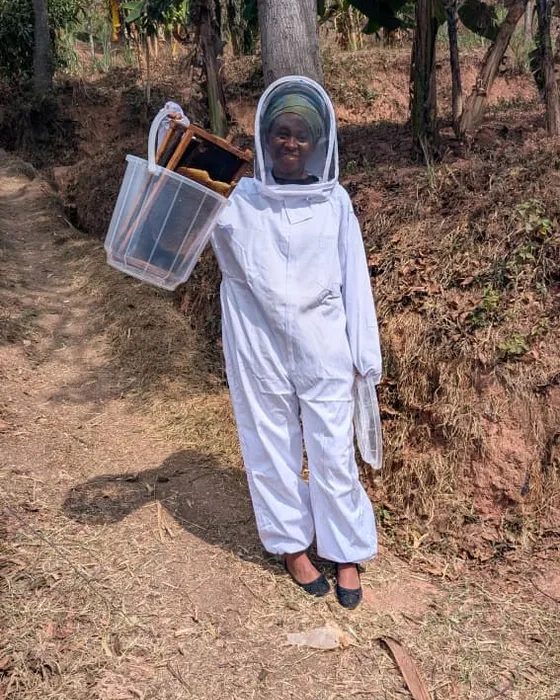 A smiling beekeeper in a white suit carrying hive frames in a basket